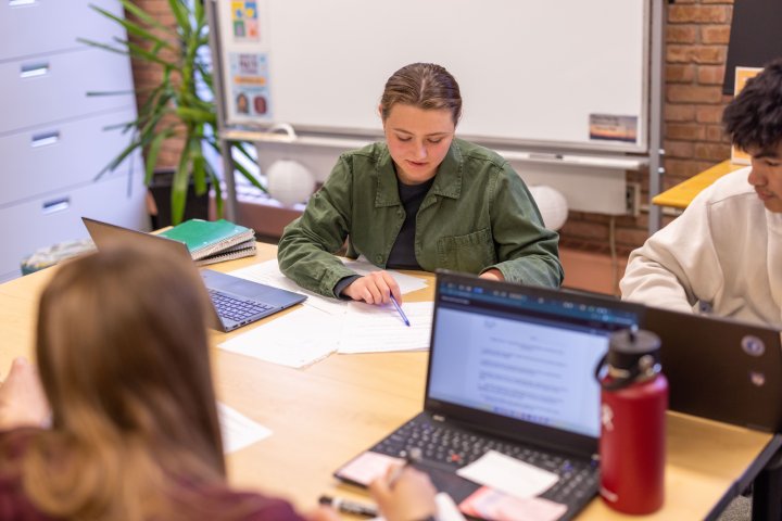 Three students work together at a table, focusing on their laptops and notes in a study area. One student in a green jacket reviews papers, while the others type on their laptops. A whiteboard and potted plant are visible in the background.